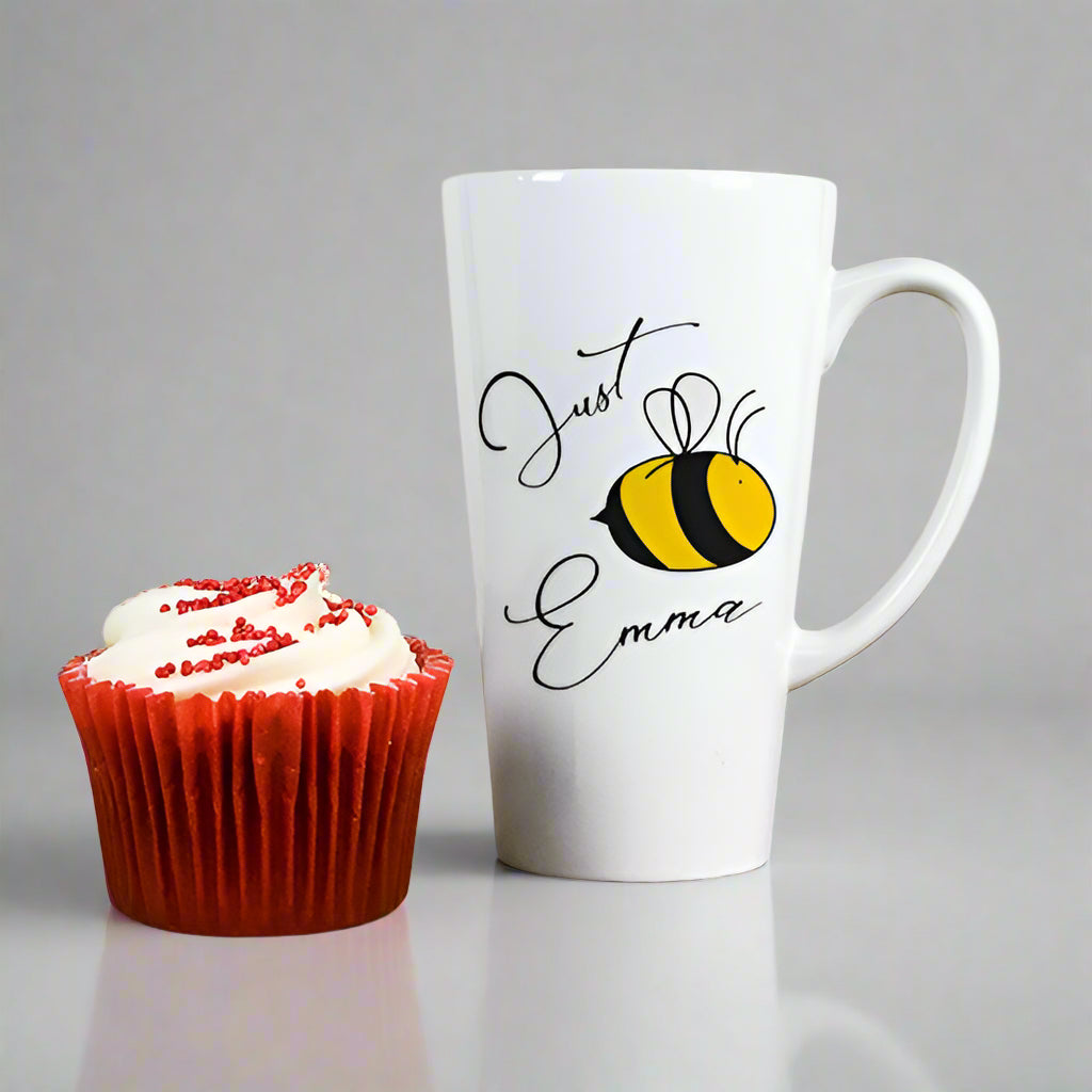 White mug with 'Just Emma' and a bee design next to a red cupcake with white frosting on a wooden surface.