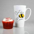 White mug with 'Just Emma' and a bee design next to a red cupcake with white frosting on a wooden surface.